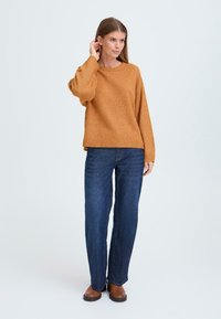 Maglione a maglia color ruggine con una vestibilità comoda e orlo a costine, abbinato a jeans a gamba dritta blu scuro e stivaletti marroni. Sfondo neutro.