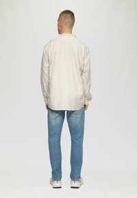 Lichtgekleurde, gestreepte blouse met lange mouwen van katoen, gecombineerd met blauwe spijkerbroek en witte sneakers, van achteren getoond.