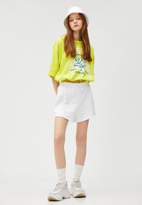Neon grøn oversized t-shirt med grafikprint, parret med hvide shorts med elastisk talje og hvide sneakers. Modellen bærer en hvid bøttehat.