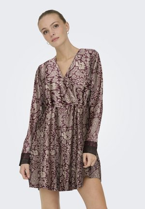 Mujer con un vestido de manga larga color granate con motivos florales y de estilo paisley en beige, de pie frente a un fondo claro y liso.