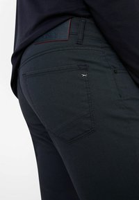 Dark blue textured pants with a smooth finish; features a leather tag with "Brax" and a small silver accent near the pocket.