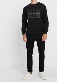 Svart bomullströja med en stor Calvin Klein Jeans-logotyp i mitten, tillsammans med svarta jeans och vita sneakers.