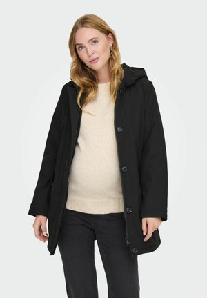Veste noire avec capuche, dotée d'une fermeture éclair et de boutons, de poches avant et d'une coupe décontractée. Portée sur un pull léger et un jean foncé.