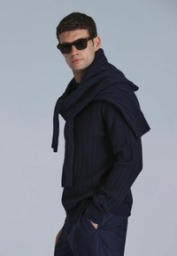 Marineblauer Kabelstrickpullover mit Schalkragen, kombiniert mit dunklen Sonnenbrillen und marineblauen Hosen, die aus strukturiertem Material gefertigt sind und einen lässigen Schnitt haben.