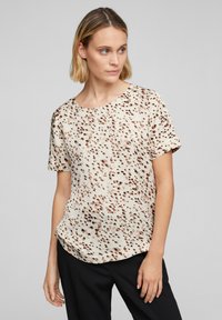 Blusa a maniche corte in crema con un motivo di pois marroni sparsi, caratterizzata da un collo rotondo e una vestibilità rilassata. Texture morbida, tessuto leggero.