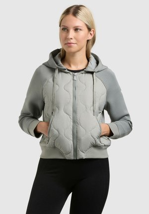 Młoda kobieta o blond włosach, ubrana w jasnoszarą pikowaną bluzę na zamek i czarne spodnie, z rękami w kieszeniach, spoglądająca lekko w prawo.