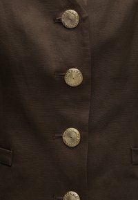 Σκούρο καφέ ύφασμα με τέσσερα περίτεχνα χρυσά κουμπιά σε κάθετο διάταξη, κάθε κουμπί ραμμένο μέσω καφέ βελονιών που ταιριάζουν.