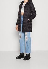 Svart puffjacka med huva, med dragkedja framtill och sidofickor; bärs med ljusblå, slitna raka jeans och svarta stövlar.