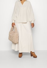 Krämfärgad oversized skjorta med vida ärmar, ihop med en lång krämfärgad kjol. Håller en texturerad beige päskväska och bär bruna klackstövlar.