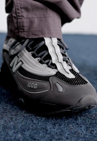 Scarpa sportiva grigia e nera realizzata con materiali in rete e sintetici, caratterizzata da una tomaia testurizzata, lacci e il marchio "1000" sul lato.