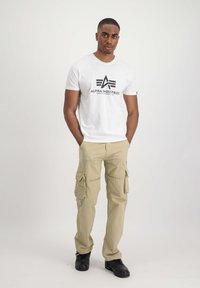 Camiseta blanca de algodón con el logo negro de "Alpha Industries", combinada con pantalones cargo beige que presentan bolsillos laterales y zapatillas negras.