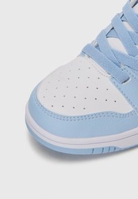 Gros plan sur l'embout d'une sneaker blanche et bleu clair avec des perforations, des détails de couture et des lacets bleu clair sur un fond blanc.