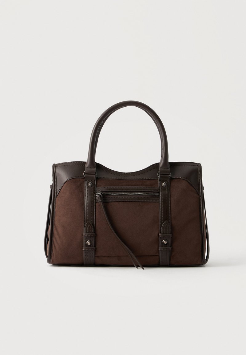 Sac à main en cuir et daim marron foncé avec deux poignées, poche avant zippée et coutures décoratives sur un fond uni clair.