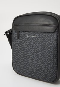 Sort skuldertaske med tekstureret stof, der har et logomønster, glat kant og en lynlåslomme foran. Logoet siger "Calvin Klein."