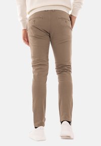 Pantaloni slim-fit in cotone marroni con due tasche posteriori, abbinati a sneaker bianche, visti da dietro. Texture liscia e design su misura.