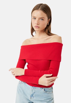 Giovane donna con capelli castani lisci indossa un maglione rosso a coste con spalle scoperte e jeans azzurri, braccia incrociate, su sfondo bianco.