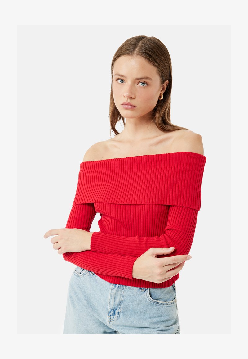 Jeune femme aux cheveux bruns et raides porte un pull côtelé rouge à épaules dénudées et un jean bleu clair, les bras croisés, sur fond blanc.