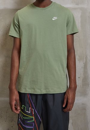 Mężczyzna w jasnooliwkowej koszulce Nike z krótkim rękawem i czarnych szortach z kolorowymi liniami logo Nike stoi przy betonowej ścianie.