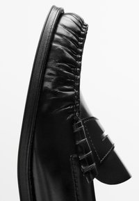 Close-up of a black leather loafer showing detailed stitching and gathered leather along the upper edge.