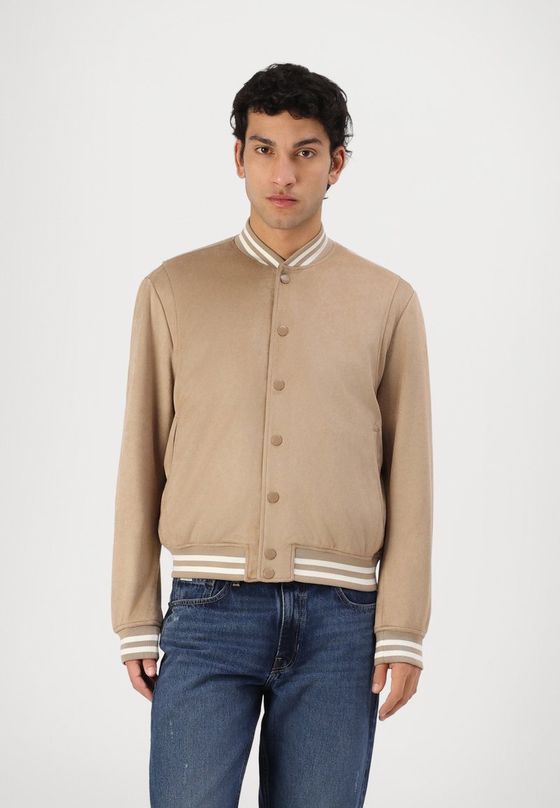 Beige bomberjakke med en jevn tekstur, med knappelukking og stripete ribbede mansjetter og kant. Benyttes over blå jeans.