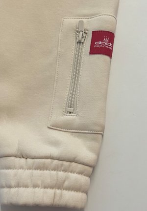 Light beige sweatshirt fabric with a zippered pocket and a red label. Cuffed sleeve with visible stitching and smooth texture.