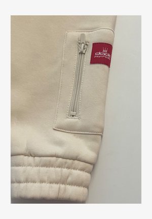 Hellbeiger Sweatshirtstoff mit einer Reißverschlusstasche und einem roten Etikett. Ärmelbündchen mit sichtbarer Naht und glatter Textur.
