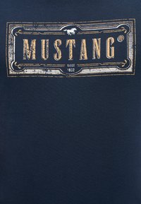 Navy blue t-shirt with a textured, cream-colored graphic featuring the word "MUSTANG" and a horse motif, established since 1932.
