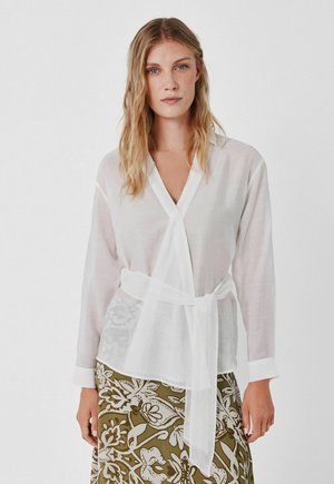 Donna che indossa una blusa bianca trasparente a portafoglio con maniche lunghe e cintura annodata in vita, abbinata a una gonna verde oliva con fantasia.