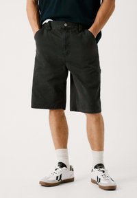 Pantalones cortos de algodón negros con un corte clásico, que cuentan con bolsillos frontales y cierre de botón, combinados con zapatillas blancas y calcetines de longitud media.
