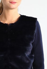 Granatowa kurtka z kołnierzem z imitacji futra, o gładkiej fakturze i okrągłym dekolcie, uzupełniona miękkim, dzianinowym korpusem.
