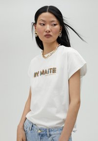 Bílá bavlněná tričko s krátkým rukávem, zdobené vícebarevným korálkovým nápisem "BY MAITE" a "SPRING SUMMER 2024" v serifovém písmu. Světle modré džíny.