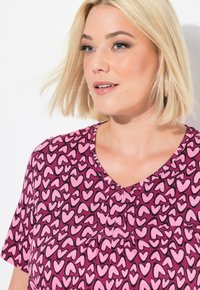 Korte mouw top in roze en paars met een hartjespatroon, voorzien van een v-hals en geplooid detail in het midden. Zachte, gladde stoftextuur.
