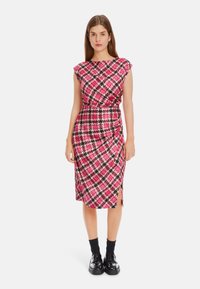 Rosa und schwarze gemusterte Kleid mit kurzen Ärmeln, seitlichem Schlitz und figurnahem Schnitt, aus strukturiertem Stoff gefertigt. Kombiniert mit schwarzen Schuhen und Socken.