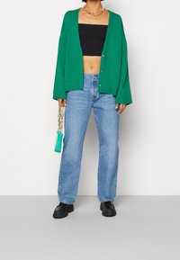 Groene oversized cardigan met lange mouwen, zwarte crop top, lichtblauwe rechttoe-rechtaan jeans en zwarte schoenen. Beschikt over een kettingaccessoire.