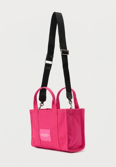 Ροζ τσάντα tote από καμβά με δύο επάνω χερούλια και αποσπώμενο μαύρο λουράκι, με λευκή ετικέτα λογότυπου στη μία πλευρά.