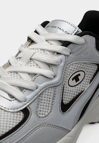 Λευκό αθλητικό παπούτσι με διχτυωτό επάνω μέρος, ασημένιες λεπτομέρειες και υφασμάτινες υφές. Διαθέτει μαύρο λογότυπο και επίπεδα λευκά κορδόνια.