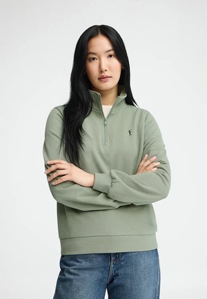 Kobieta z długimi czarnymi włosami, ubrana w zieloną bluzę z zamkiem do połowy długości i niebieskie jeansy, stojąca z założonymi ramionami na tle białego tła.