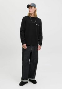 Black sweatshirt with a crew neck, gray cap, and oversized dark denim jeans with rolled cuffs. Features chain and tattooed hands.