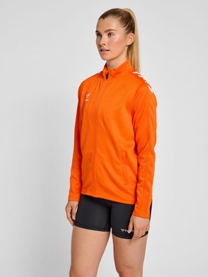 Ung kvinde med blond hestehale iført en lyse orange sport jakke med lynlås og sorte atletik shorts, stående mod en ensfarvet baggrund.