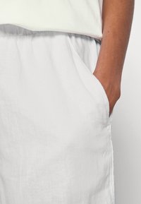 Personne à la peau foncée portant un pantalon blanc ample avec une taille élastique, la main placée de manière décontractée dans la poche droite.