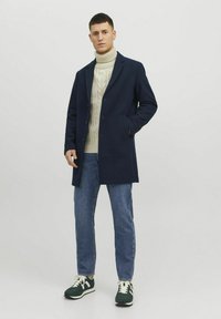 Marinblå ullrock med slagkrage, enfärgad design, kombinerad med en krämfärgad kabelstickad polotröja, blå jeans och mörkgröna sneakers.