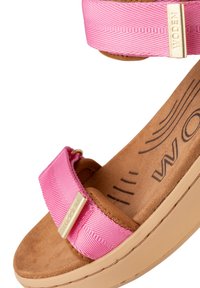 Pink strap sandal featuring textured fabric, golden hardware, and a cushioned brown footbed with a wavy pattern. Lightweight tan sole.