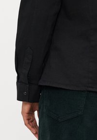Zwarte shirt met knoopmanchetten, voorzien van een puntige stofplacket en gladde textuur. Draag het over groene corduroy broek met ruitvormige zakdetails.