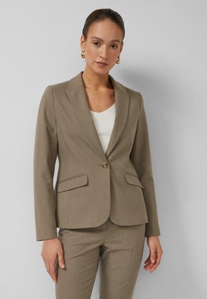 Kobieta w beżowej, dopasowanej marynarce i dopasowanych spodniach oraz złotych kołowych kolczykach, stojąca na tle jednolitego jasnoszarego tła.