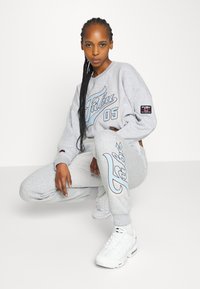 Šedá mikina a odpovídající tepláky s grafikou „FUBU 05“ v modré barvě. Tepláky mají postranní logo. Model má na sobě bílé tenisky.