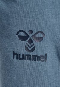 Blå atletisk tyg med en texturerad yta, med en framträdande svart logotyp av två abstrakta vingar och texten "hummel" nedan.