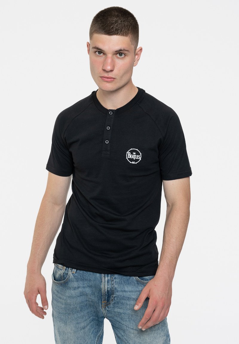 Black short-sleeve henley shirt with three buttons and a white "The Beatles" logo on the left chest. Worn with light blue jeans.