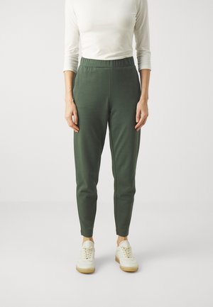 Persona vistiendo pantalones ajustados de color verde oscuro con cintura elástica, camisa blanca de manga larga y zapatillas blancas, de pie sobre un fondo liso.