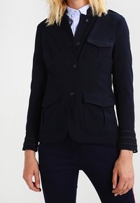 Giacca blu navy con un design aderente, dotata di quattro tasche, chiusure a bottone e dettagli testurizzati sulle maniche. Materiale liscio.