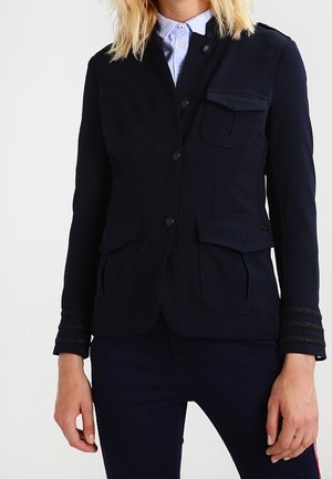 Marineblauwe jas met een getailleerd ontwerp, voorzien van vier zakken, knoopsluitingen en textuuraccenten op de mouwen. Vloeiend materiaal.